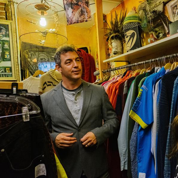 A man talks to an audience member in the back of a used clothing store.
