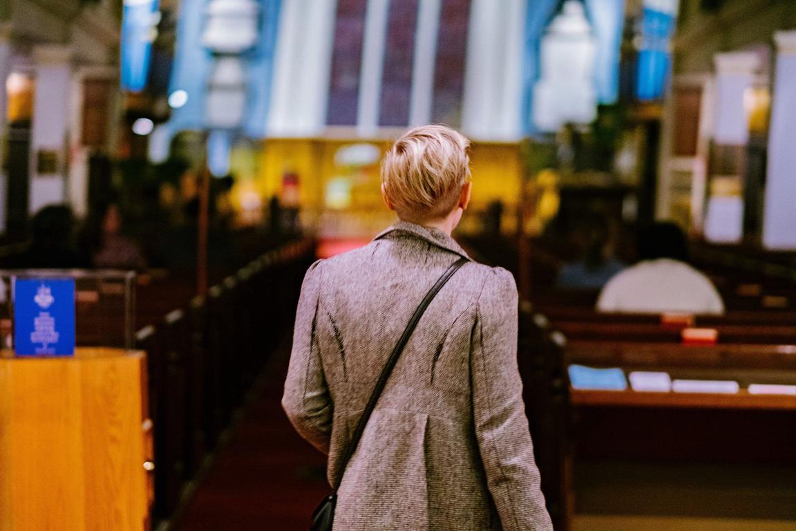 A person with short blonde hair in a grey coat with a black bag over their shoulder looks towards the alter of a church with dark wood pews.